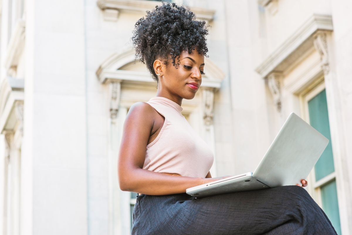 Way to Success. Young African American woman with afro hairstyle wearing sleeveless light color top, sitting by vintage office building in New York, looking down, reading, working on laptop computer.
